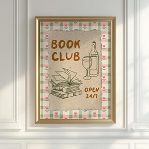 May include: A framed print with a gingham background in pink and green. The print features a hand-drawn illustration of a stack of books, a wine glass, and a bottle of wine. The text "BOOK CLUB OPEN 24/7" is written in brown and green.