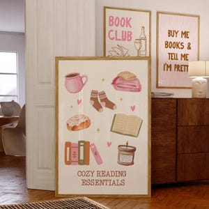 May include: A printable poster with a beige background and pink accents. The poster features illustrations of cozy reading essentials, including a book, a cup of coffee, a candle, a blanket, a cinnamon roll, and a stack of books. The text "Cozy Reading Essentials" is at the bottom of the poster.