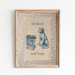 May include: A framed print with a beige background and blue floral border. The print features a black and white illustration of a dog sitting next to a stack of books. The text "booked and busy" is printed below the illustration.
