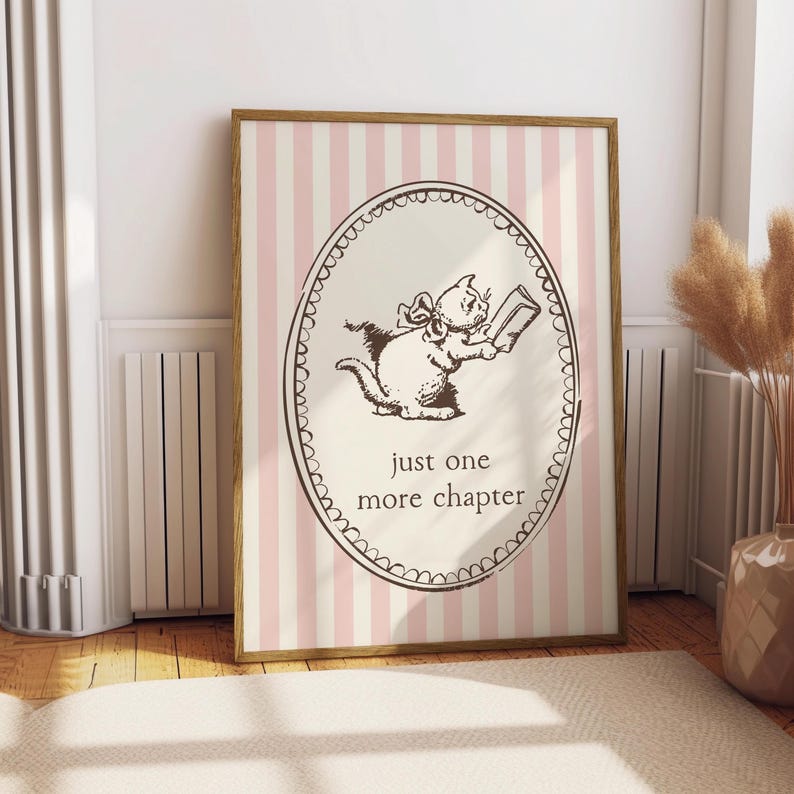 May include: A framed print with a cartoon cat reading a book, with the text "just one more chapter." The artwork is set against a pink and white striped background, framed in a light brown wood.