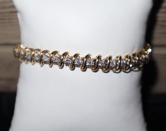 May include: A gold bracelet featuring a series of clear, sparkling stones set in a curved design. The bracelet is displayed on a white cushion, highlighting its elegant and timeless appeal. A classic piece of jewellery.