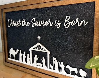 May include: Wooden framed sign with the text "Christ the Savior is Born" in white script. The sign features a white silhouette of the nativity scene against a black background. The frame is a natural wood color.