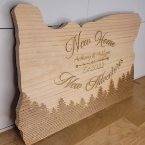 May include: Wooden sign in the shape of Oregon, engraved with the words "New Home, New Adventures" and the names "Anthony & Ashlynn, Est 2022." A forest silhouette is carved at the bottom. The sign is made of light-colored wood.
