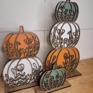 Può includere: Decorazioni a forma di zucca in legno arancione, bianco e verde, con intricati ritagli floreali. Le zucche sono impilate e disposte su basi di legno, creando un'esposizione a tema autunnale. Le zucche sono di varie dimensioni.