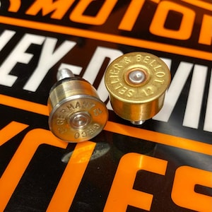 Puede incluir: Dos tapones de botella con forma de cartucho de escopeta de color latón. Un tapón está marcado "GERMANY 12 GECO" y el otro "SELLIER & BELLOT 12". Los tapones tienen un poste plateado.