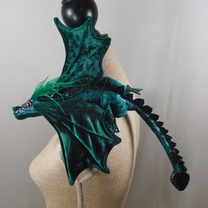 May include: A handcrafted, emerald green dragon sculpture with velvet wings and a textured body. The dragon features red eyes, green feather accents, and a long, segmented tail. The sculpture is designed to be worn as a shoulder piece.