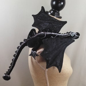 May include: A black dragon shoulder accessory with detailed wings and a long tail. The dragon has a silver spine and accents, with green eyes. The accessory is designed to be worn on the shoulder, adding a fantasy element to any outfit.