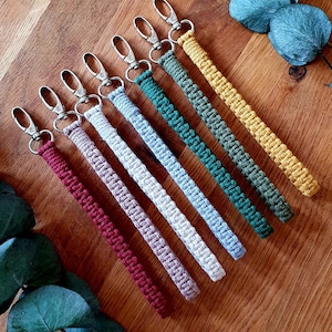 Op de afbeelding: Acht kleurrijke gebreide sleutelhangers met zilveren sleutelringen. De sleutelhangers zijn in een rij op een houten oppervlak gerangschikt. De kleuren zijn rood, roze, wit, lichtblauw, donkerblauw, groen en geel.