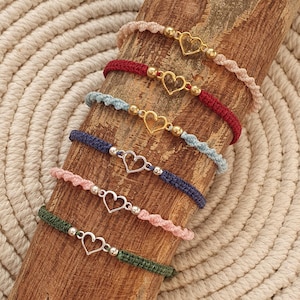 Op de afbeelding: Zes kleurrijke vriendschapsarmbanden met gouden en zilveren hartcharms. De armbanden zijn gemaakt van verschillende gekleurde koorden en zijn op een houten oppervlak gelegd.