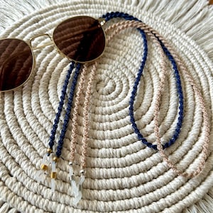 May include: A pair of sunglasses with round brown lenses and a gold-tone frame, attached to a dual-strand eyeglass chain. The chain features one strand of dark blue braided cord and one strand of beige braided cord, finished with metal beads.