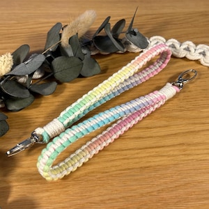 Op de afbeelding: Twee macramé polsbandjes in pastel regenboogkleuren. Elk polsbandje heeft een witte lus en een zilverkleurige metalen sluiting. De polsbandjes zijn gemaakt van geweven katoenen koord en zijn geschikt voor sleutels of kleine accessoires.