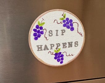 May include: A round white embroidery hoop with the text "Sip Happens" in gray lettering. Three bunches of purple grapes with green stems are embroidered on the hoop.