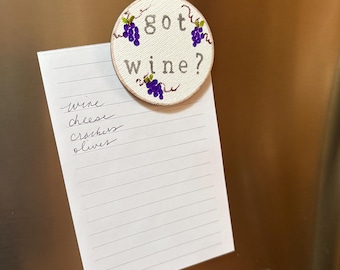 May include: A white round magnet with the text "got wine?" and a grapevine design. The magnet is attached to a white notepad with a handwritten grocery list that includes "wine, cheese, crackers, olives".