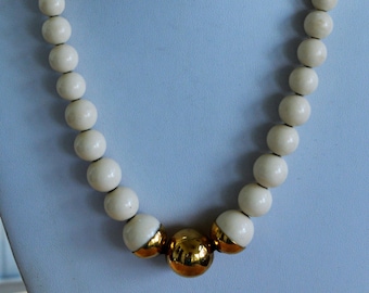 May include: Cream-colored beaded necklace with a gold-colored accent. The necklace features large, round beads and a central cluster of gold-toned beads. The necklace is displayed on a light blue mannequin.