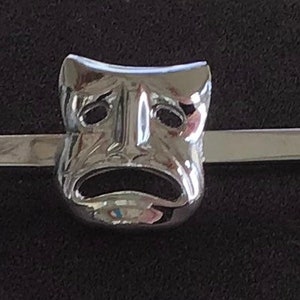 May include: A silver tie bar with a stylized, sad-faced mask design.