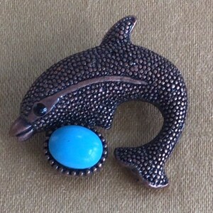May include: A bronze-colored dolphin-shaped brooch with a textured surface. The dolphin has a turquoise-colored, oval-shaped stone set in a bronze bezel. The brooch has a pin clasp on the back.