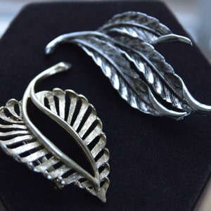 Pode incluir: Dois broches de prata em forma de folha. Um broche é uma única folha com um design detalhado. O outro broche é de duas folhas com um design mais simples.