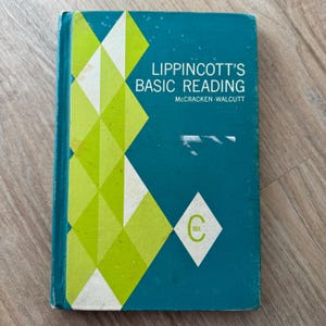 May include: A teal blue book with a green and white geometric design on the cover. The title is "Lippincott's Basic Reading" with the author's name "McCracken-Walcutt" below. The book is labeled "Book C".