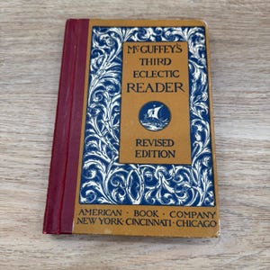 May include: A vintage, gold and blue book titled "McGuffey's Third Eclectic Reader, Revised Edition" with a decorative border and a ship illustration. The book is published by the American Book Company, New York, Cincinnati, Chicago.