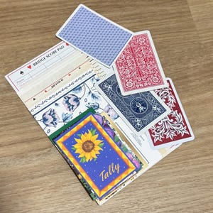May include: A collection of playing cards and stationery. The cards feature red and blue floral designs, and a blue patterned back. Stationery includes a bridge score pad and a card with a sunflower and the word "Tally".