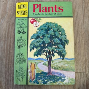 Pode incluir: Capa de livro vintage "Living Science Plants". A capa apresenta uma grande árvore verde com uma estrada sinuosa, uma maçã e uma espiga de milho. O título "Plants" está em letras vermelhas grandes, com a legenda "A preface to the study of plants."