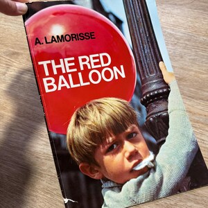 May include: A book cover showing a young boy with fair hair and a red balloon. The title "THE RED BALLOON" is in white letters against the red balloon. The author's name, A. LAMORISSE, is above the title. The boy is holding onto a lamppost.