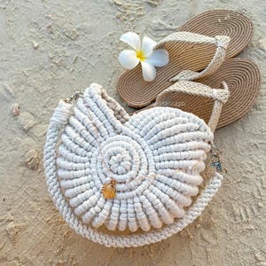 Puede incluir: Un bolso blanco de macramé con forma de concha marina con un collar con colgante de concha dorada, sandalias de cuerda beige y una flor blanca sobre una superficie arenosa. El bolso tiene una larga correa de cuerda y herrajes plateados.