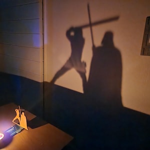 May include: A shadow of two figures holding lightsabers projected on a wall. The figures are facing each other in a fighting stance. The shadow is cast by a wooden cutout of the figures.