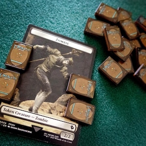 May include: A rectangular trading card featuring a zombie illustration with the text Zombie and Token Creature - Zombie, surrounded by several small brown rectangular tokens with the Dragon Shield logo printed on their surfaces.