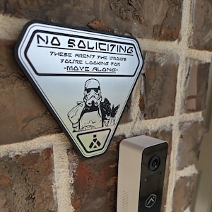 Könnte beinhalten: Ein silbernes und schwarzes Metallschild mit der Aufschrift "NO SOLICITING" und einem Star Wars-Thema. Das Schild zeigt einen Sturmtruppler und den Satz "THESE AREN'T THE DROIDS YOU'RE LOOKING FOR - MOVE ALONG-". Eine weiße und schwarze Türklingel ist darunter montiert.