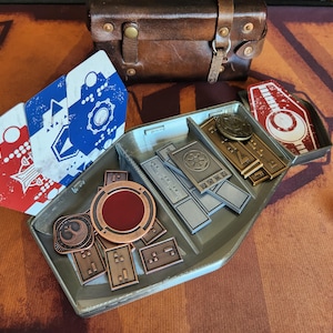 May include: A grey plastic tray with compartments for storing small items. The tray contains a variety of metal tokens, including a red and gold token with a symbol, a silver token with a symbol, and several smaller gold tokens. There are also two cards with a red, white and blue design.