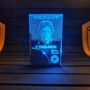 Puede incluir: Placa rectangular de acrílico azul brillante con un retrato de Han Solo. La placa incluye texto en inglés y otro idioma, junto con un símbolo. El fondo es oscuro, con dos formas amarillas a cada lado.
