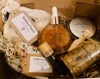 May include: A gift box containing a Golden Touch herbal cleansing sponge, a bottle of She Who Glows body oil, a tin of She Glows dusting powder with a wooden scoop, a floral fabric bag labeled Golden Glow, and a jar of Golden Soak bath salts.