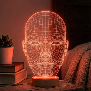 Puede incluir: Lámpara moderna con forma de rostro geométrico, con un cálido resplandor naranja. La lámpara presenta un diseño de estructura de alambre 3D de un rostro humano, montada sobre una base de madera. La lámpara está enchufada y sobre una superficie de madera.
