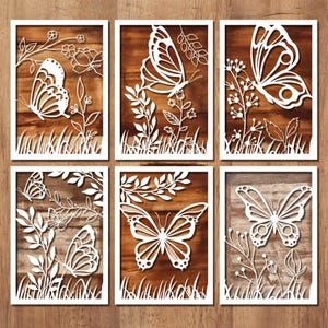 Könnte beinhalten: Sechs gerahmte Holzpaneele mit weißen, lasergeschnittenen Schmetterlings- und Blumendesigns. Die Schmetterlinge und Pflanzen sind vor einem braunen Holzmaserungshintergrund angeordnet. Jedes Paneel hat einen weißen Rahmen, wodurch ein zusammenhängendes Wandkunst-Set entsteht.