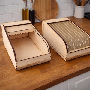 May include: Two wooden bread boxes with a natural finish sit on a wooden countertop. One box is open, revealing an empty interior. The other box has a slatted, roll-top lid. Both boxes have dark brown trim and are made of light-colored wood.
