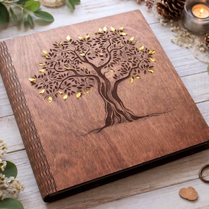 May include: A wooden photo album with a tree design. The cover is made of dark brown wood and features a detailed tree engraving with gold-colored accents. The album is rectangular and appears to be closed. The overall aesthetic is rustic and elegant.