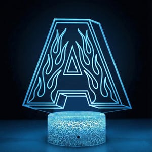 May include: A neon blue letter "A" shaped lamp with flame accents. The lamp has a geometric design and sits on a textured, circular base. The background is dark, highlighting the bright blue light.