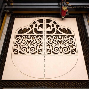 May include: A laser cutter precisely cuts a light-colored wooden sheet into a decorative, ornate design. The design is divided into four sections with intricate scrollwork and a curved bottom edge, suggesting a potential box or container.