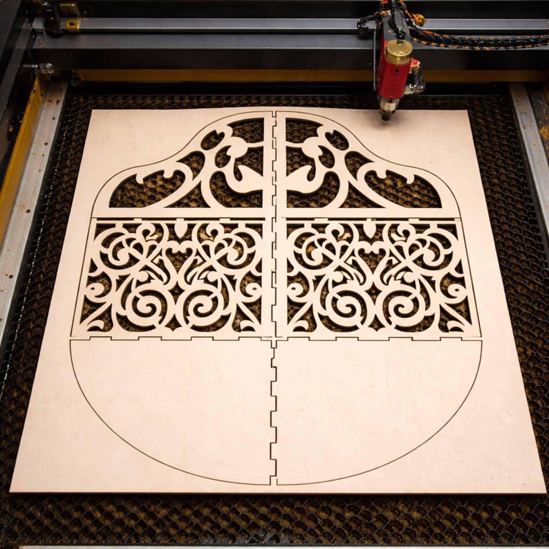 May include: A laser cutter precisely cuts a light-colored wooden sheet into a decorative, ornate design. The design is divided into four sections with intricate scrollwork and a curved bottom edge, suggesting a potential box or container.