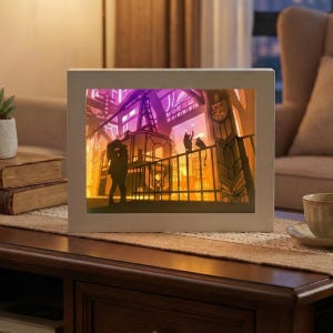 May include: A framed, illuminated art piece showing a romantic cityscape scene. Silhouetted figures of a couple kissing and cats on a balcony are set against a gradient of purple, orange, and yellow hues. The frame is a neutral off-white colour.