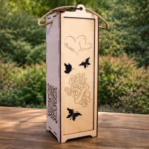 Puede incluir: Un portabotellas de vino de madera clara con asa de cuerda. La parte delantera presenta diseños grabados de corazones, mariposas y un ramo de rosas. Un patrón decorativo calado está en el lateral.