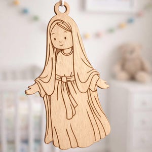 May include: Wooden cutout of a figure with long hair and flowing robes, hanging from a white string. The figure has outstretched arms and a gentle expression. The natural wood grain is visible. A decorative item for a nursery or child's room.