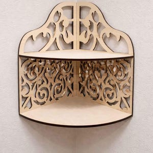 May include: A wooden corner shelf with an ornate, openwork design. The shelf has two tiers and a decorative top with a curved shape. The edges are dark brown, and the shelf is made of light-colored wood.