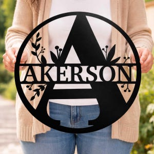 May include: A black metal family name sign with the name "AKERSON" and the letter "A" in a circular design. The sign features floral accents and is held by a person.
