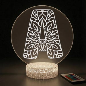 May include: A decorative LED lamp featuring the letter "A" with a floral design. The letter is white and set against a clear, circular background. The lamp sits on a cracked-patterned base and includes a remote control.