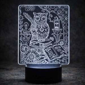 May include: A decorative LED night light featuring an owl with a feather, perched atop books. The design includes floral and celestial elements, with the word "INK" visible. The light sits on a black base.