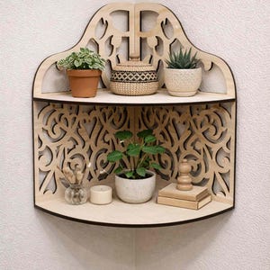 Op de afbeelding: Houten hoekplank met ingewikkelde uitgesneden patronen, met potplanten, een geweven mand en decoratieve items. De plank heeft twee niveaus en is aan een witte muur bevestigd. De plank is ongeveer 61 cm breed en 61 cm hoog.