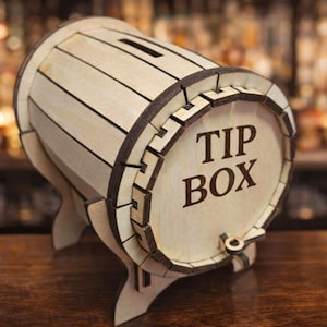 Op de afbeelding: Een houten fooienbox in de vorm van een vat, met de tekst "TIP BOX". De doos is gemaakt van lichtgekleurd hout met donkerbruine accenten en heeft een gleuf aan de bovenkant voor het plaatsen van fooien.