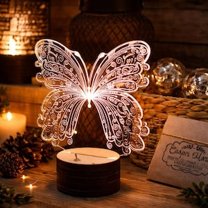 May include: An illuminated butterfly-shaped LED lamp with detailed etched patterns. The clear acrylic butterfly sits on a dark brown wooden base. The lamp emits a warm light. The lamp is displayed on a wooden surface, with other decorative items in the background.